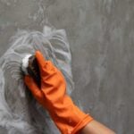 Hand in an orange glove with a scrub brush scrubbing a grey, dirty wall.