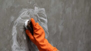 Hand in an orange glove with a scrub brush scrubbing a grey, dirty wall.
