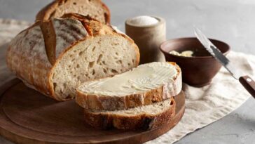loaf of rustic bread sliced on a cutting board with a wooden bowl of butter beside it