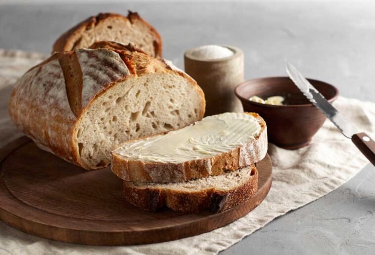 loaf of rustic bread sliced on a cutting board with a wooden bowl of butter beside it