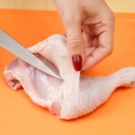 hand holding back the sking on a chicken leg quarter while it is being cut away with a knife.
