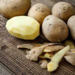 Raw potatoes on a wooden table with one peeled with the peels laying on the table in front of it.