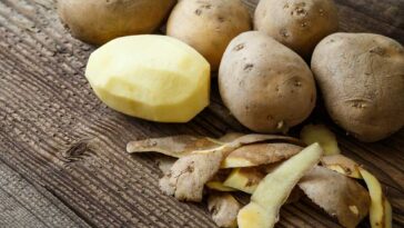 Raw potatoes on a wooden table with one peeled with the peels laying on the table in front of it.