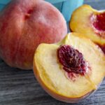 A peach cut in half with the pit exposed next to a whole peach on a wooden table