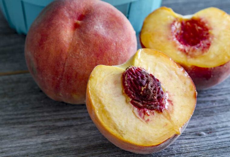 A peach cut in half with the pit exposed next to a whole peach on a wooden table