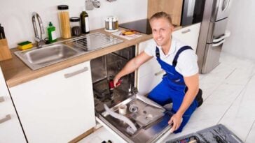 Man in overalls kneeling in front of a dishwasher he is installing