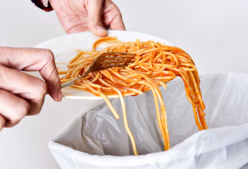 Spaghetti being scraped from a plate with a white fork into a garbage can lined with a white bag.