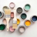 Small, open cans of paint in different colors with 2 paintbrushes laying on top on a white surfaceg