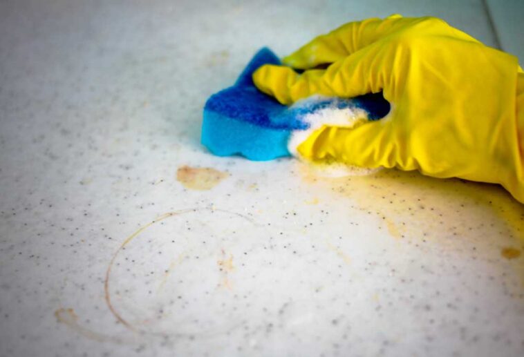 Yellow gloved hand scrubbing stains from a countertop with a blue sponge