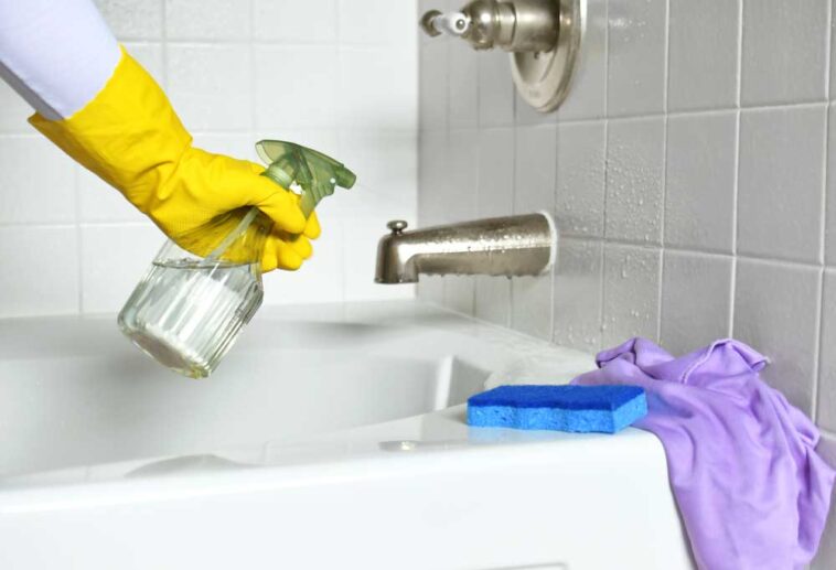 Human hand in a yellow glove using spray bottle to clean a bathtub