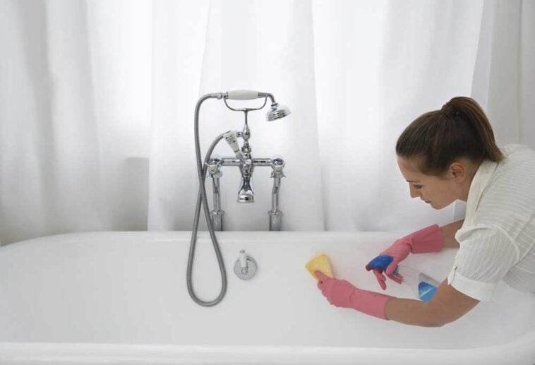 Woman leaning into white bathtub with cleaner and a rag to clean the tub.