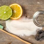 Spilled baking soda, a toothbrush, a lemon and lime slice, an empty jar and steel wool on a wooden surface