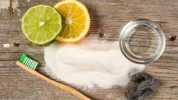 Spilled baking soda, a toothbrush, a lemon and lime slice, an empty jar and steel wool on a wooden surface