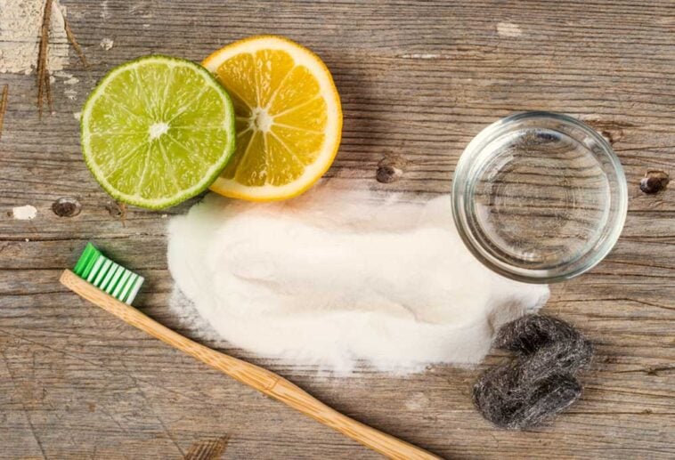 Spilled baking soda, a toothbrush, a lemon and lime slice, an empty jar and steel wool on a wooden surface